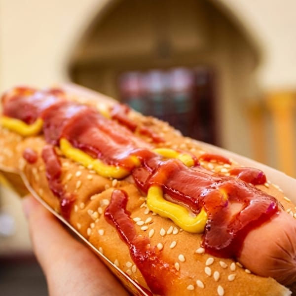 A hand holding a hot dog with ketchup and mustard on a sesame seed bun.