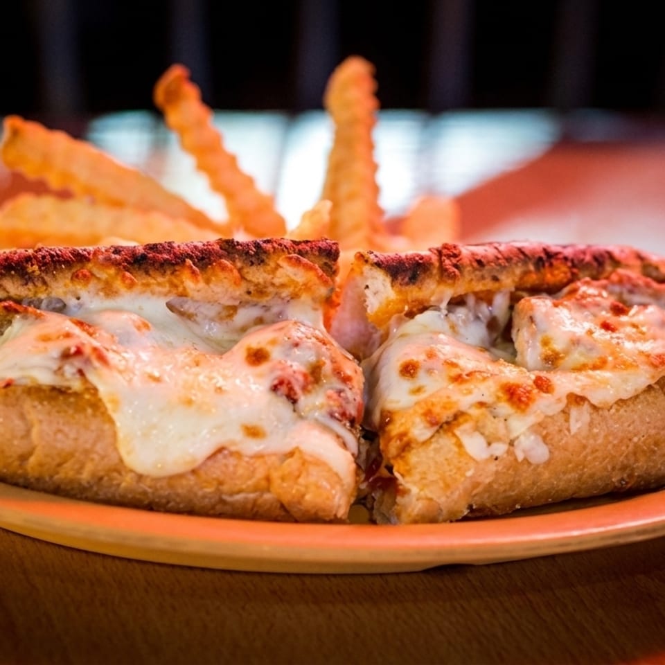 Two cheesy meatball subs on a plate with crinkle-cut fries on a wooden table.