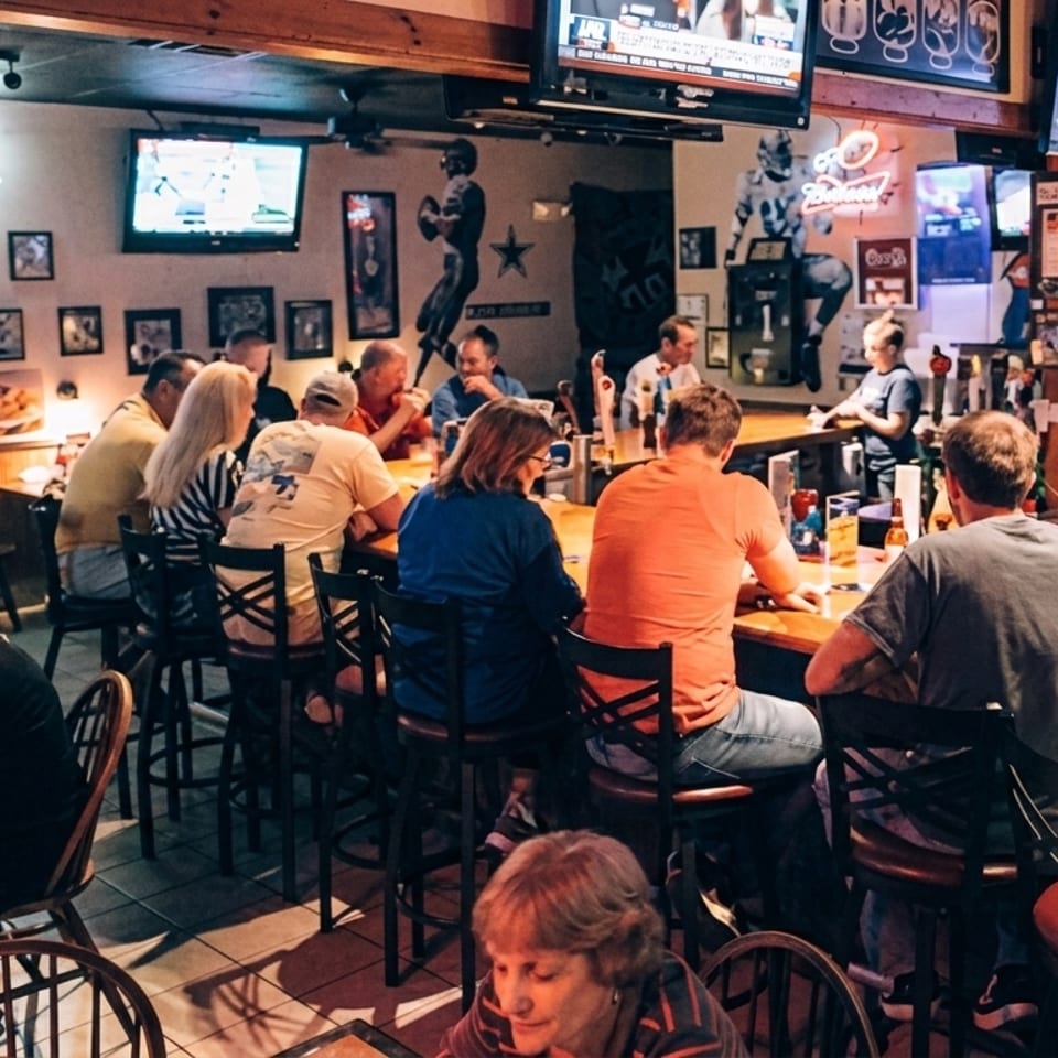 Patrons seated at a lively bar and tables in a sports-themed restaurant with multiple TVs.